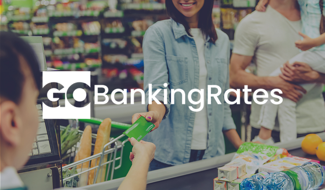 A smiling woman checking out at a grocery store with food items in a cart, featuring the GOBankingRates logo across the image.