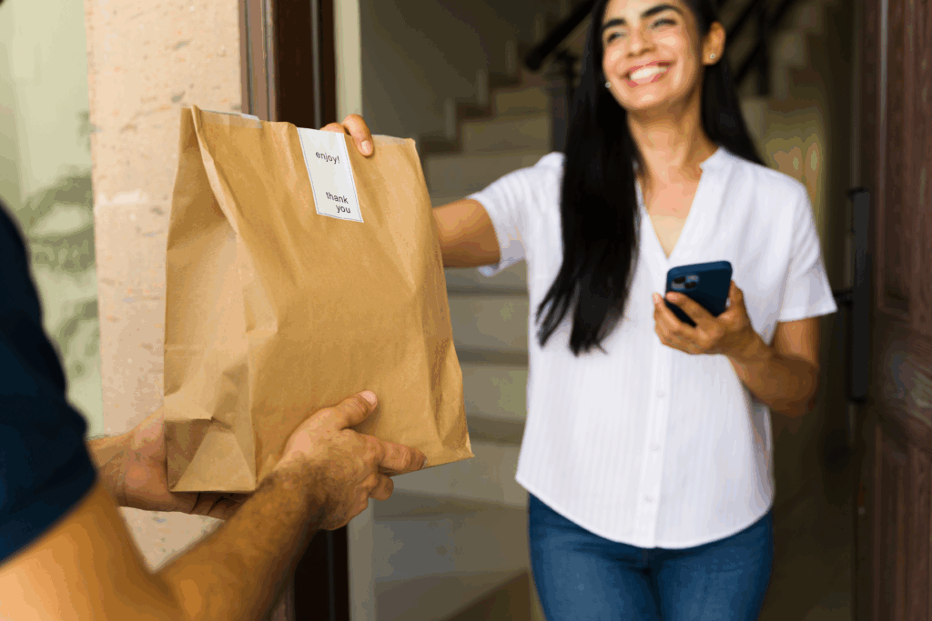 A smiling woman stands at her doorway, accepting a paper bag of food delivery from a courier while holding her phone.