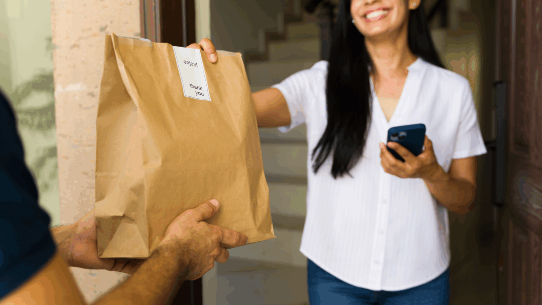 A smiling woman stands at her doorway, accepting a paper bag of food delivery from a courier while holding her phone.