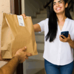 A smiling woman stands at her doorway, accepting a paper bag of food delivery from a courier while holding her phone.
