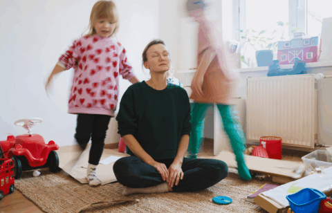 Mother sitting on the floor with eyes closed, trying to relax while children play around her in a messy room.