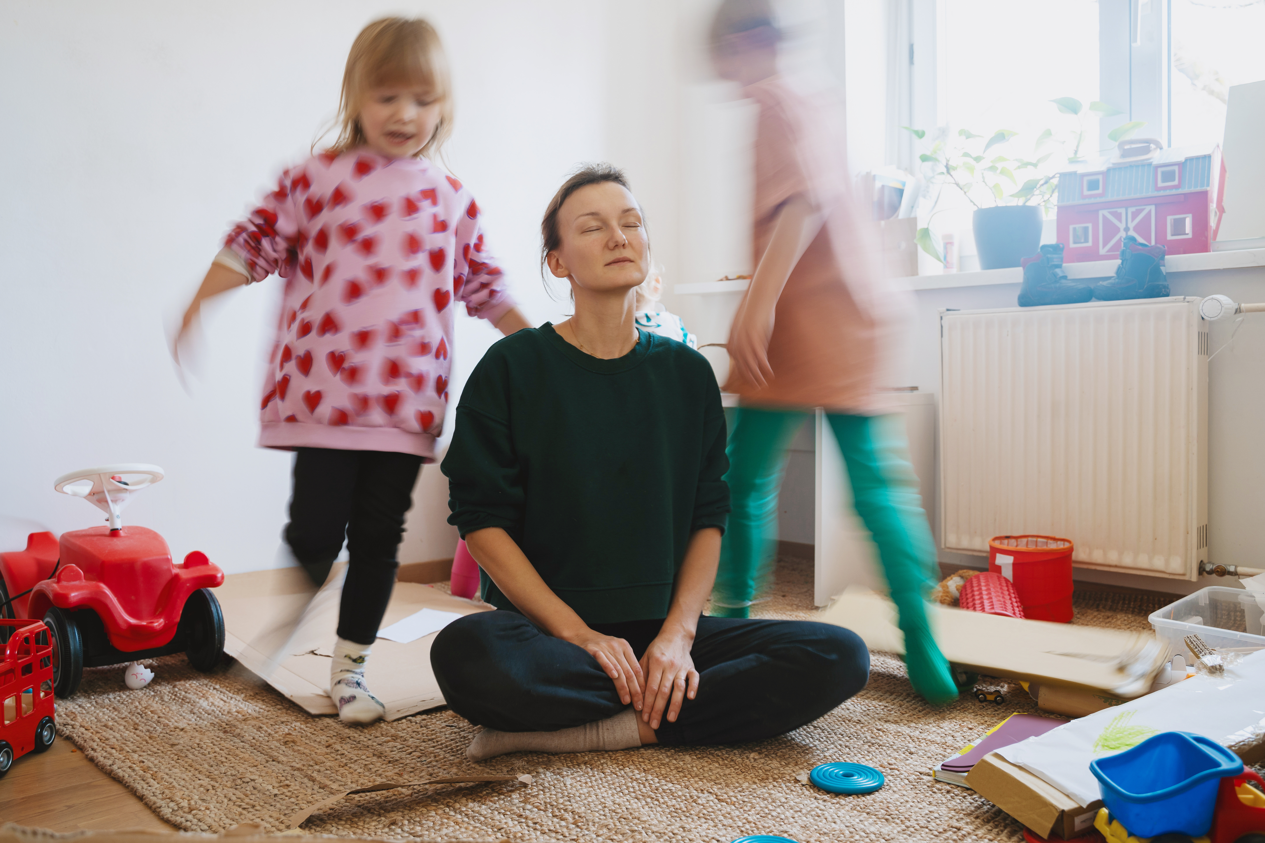Mother sitting on the floor with eyes closed, trying to relax while children play around her in a messy room.