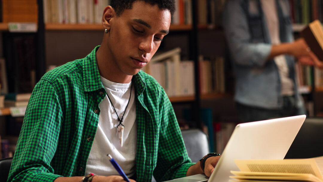 Student studying with a laptop and notebook in a library.