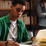 Student studying with a laptop and notebook in a library.