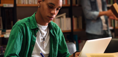 Student studying with a laptop and notebook in a library.