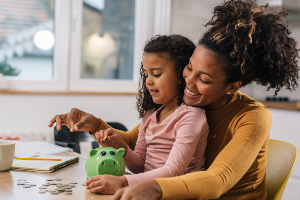 A mother and daughter smile while putting coins into a green piggy bank at the kitchen table.