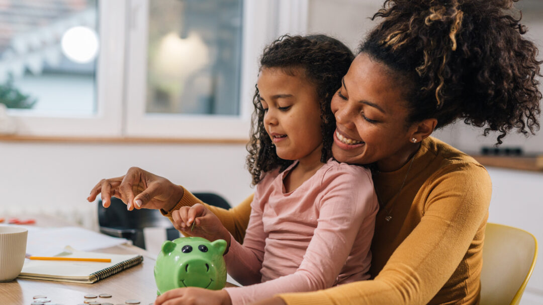 A mother and daughter smile while putting coins into a green piggy bank at the kitchen table.