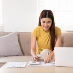 Woman using a calculator and laptop to review monthly budget at home.