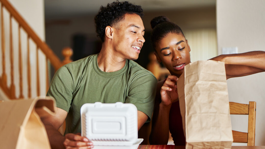 A couple sits at a dining table enjoying takeout food together at home.