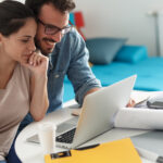 A couple sits together at a table reviewing financial documents and plans on a laptop.