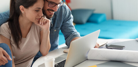 A couple sits together at a table reviewing financial documents and plans on a laptop.