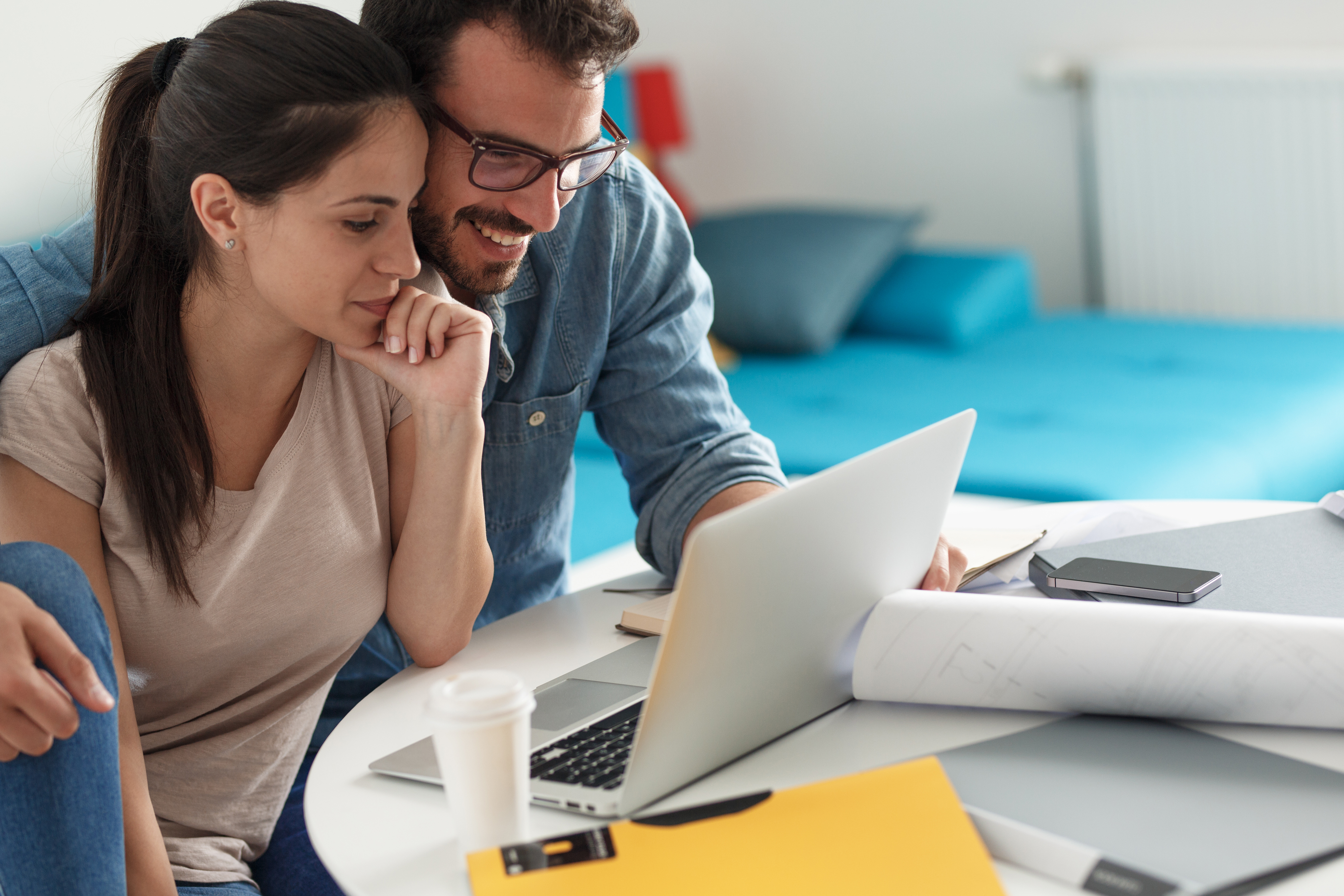 A couple sits together at a table reviewing financial documents and plans on a laptop.