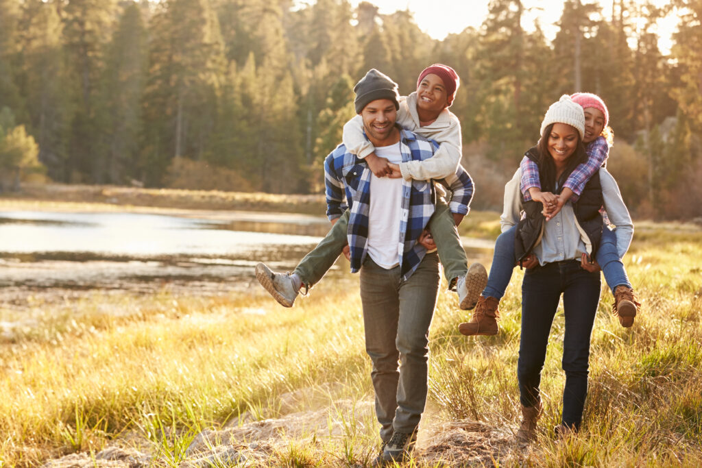 Happy family walking outdoors by a lake, with parents giving their children piggyback rides.