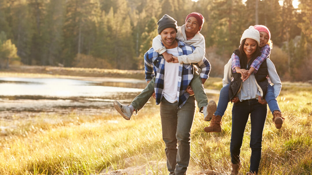 Happy family walking outdoors by a lake, with parents giving their children piggyback rides.