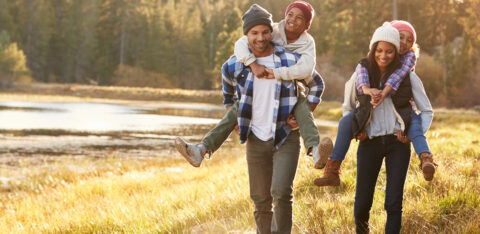Happy family walking outdoors by a lake, with parents giving their children piggyback rides.