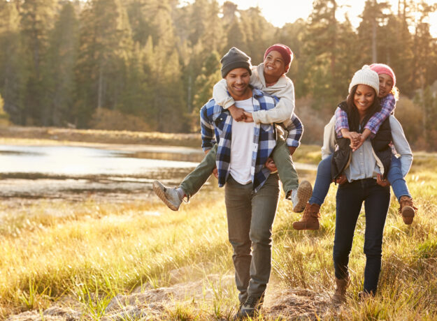 Happy family walking outdoors by a lake, with parents giving their children piggyback rides.