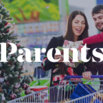 A smiling family shopping for holiday gifts, standing by a decorated Christmas tree with a cart full of presents. The Parents logo is overlayed.