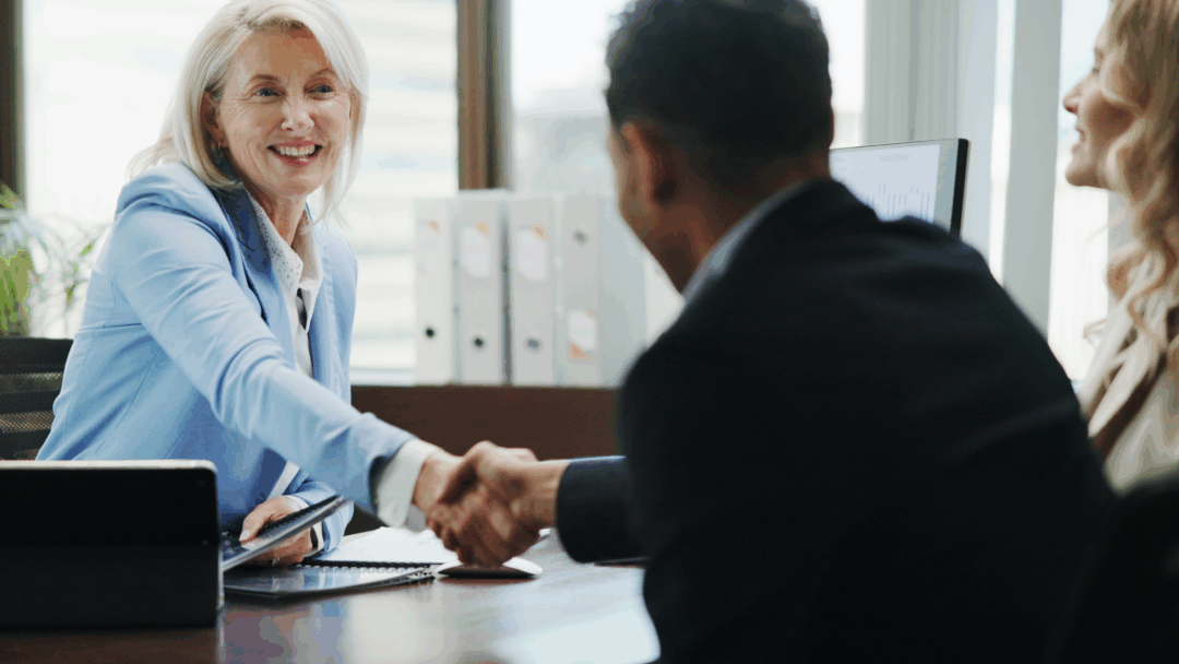 Smiling businesswoman in a meeting shaking hands with a client across a desk.