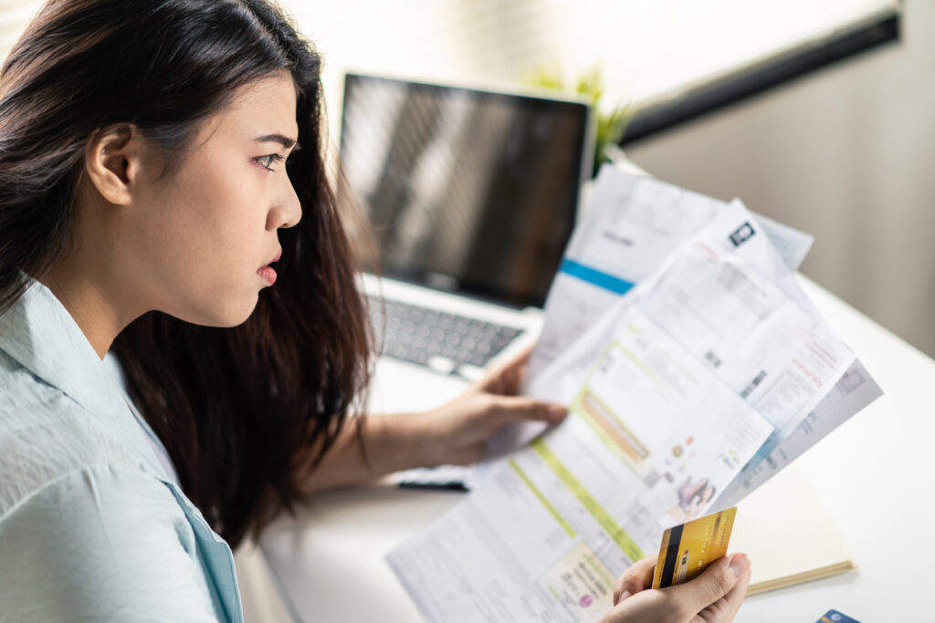 A woman sits at her desk holding several bills and a credit card, looking worried as she reviews her finances in front of a laptop.