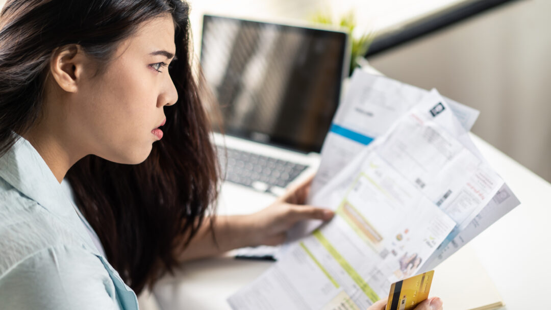 A woman sits at her desk holding several bills and a credit card, looking worried as she reviews her finances in front of a laptop.