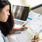 A woman sits at her desk holding several bills and a credit card, looking worried as she reviews her finances in front of a laptop.