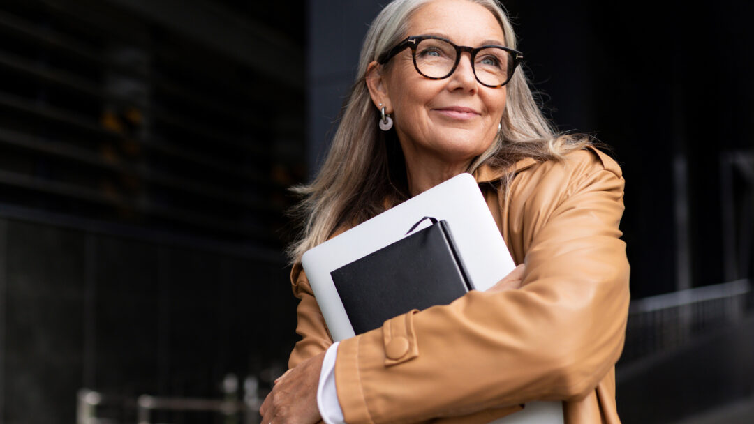 Confident older woman with gray hair smiling while holding a laptop and notebook outdoors.