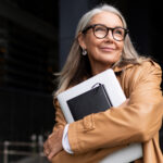 Confident older woman with gray hair smiling while holding a laptop and notebook outdoors.