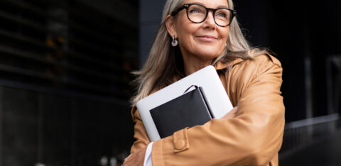 Confident older woman with gray hair smiling while holding a laptop and notebook outdoors.