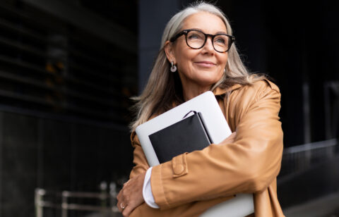 Confident older woman with gray hair smiling while holding a laptop and notebook outdoors.