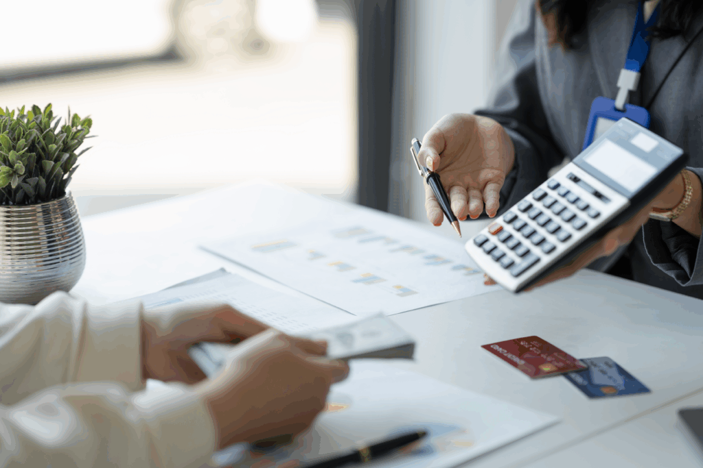 A person holds a calculator and pen while discussing paperwork with someone holding cash at a desk.