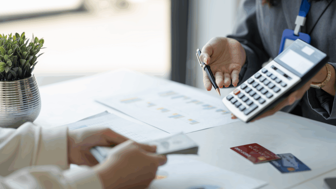 A person holds a calculator and pen while discussing paperwork with someone holding cash at a desk.