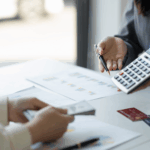 A person holds a calculator and pen while discussing paperwork with someone holding cash at a desk.