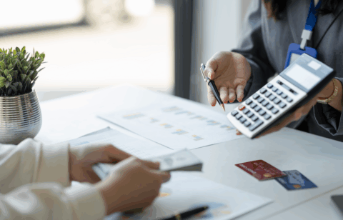 A person holds a calculator and pen while discussing paperwork with someone holding cash at a desk.
