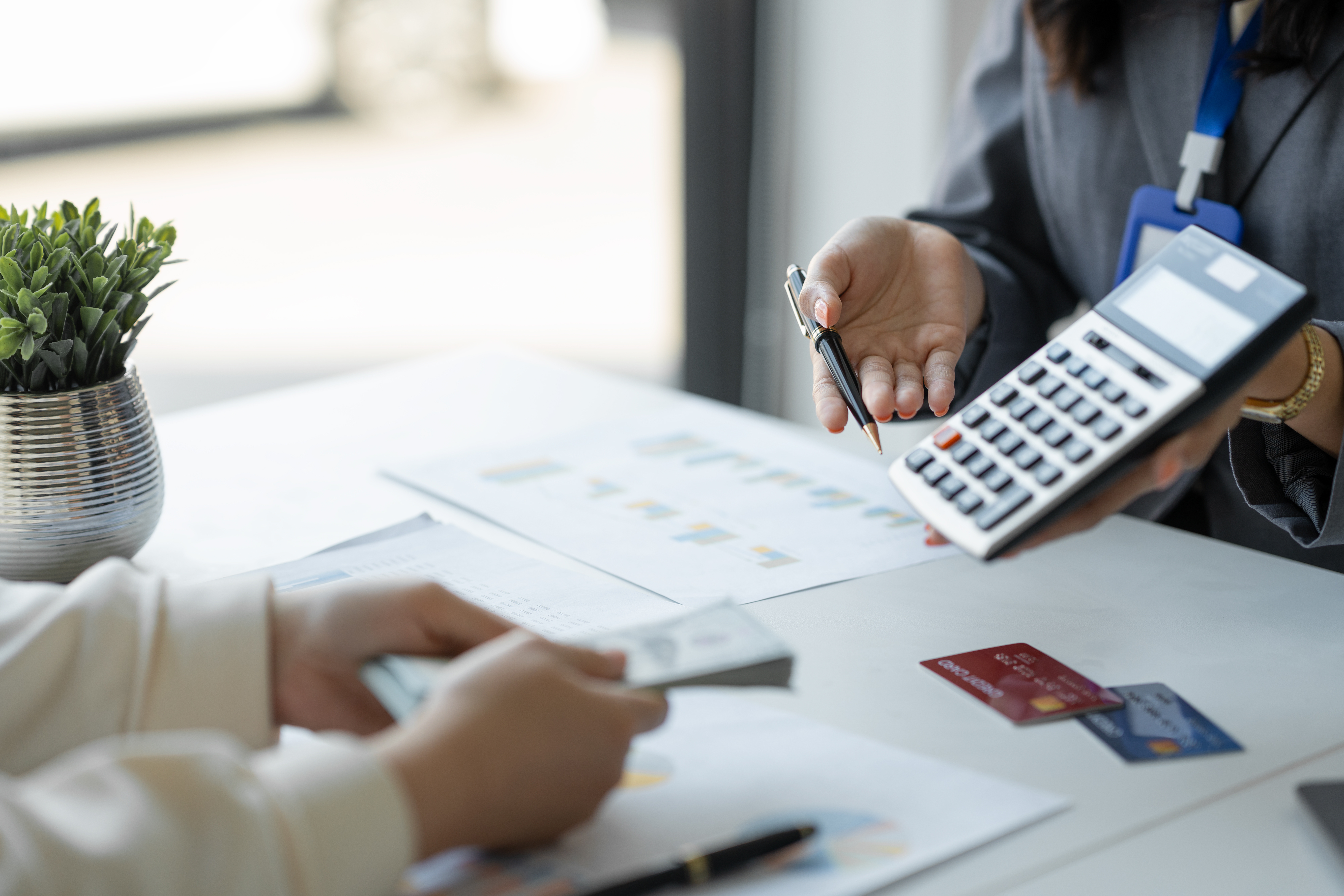A person holds a calculator and pen while discussing paperwork with someone holding cash at a desk.