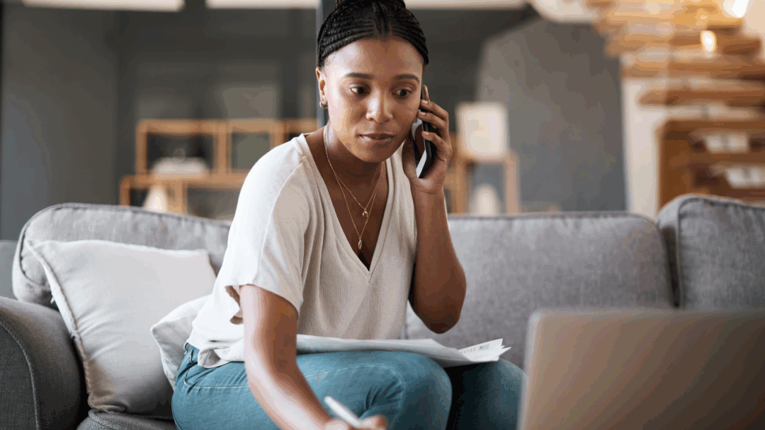 A woman sitting on a couch talks on the phone while reviewing papers and taking notes on her laptop.