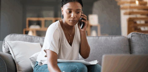 A woman sitting on a couch talks on the phone while reviewing papers and taking notes on her laptop.