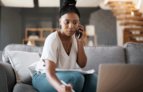 A woman sitting on a couch talks on the phone while reviewing papers and taking notes on her laptop.