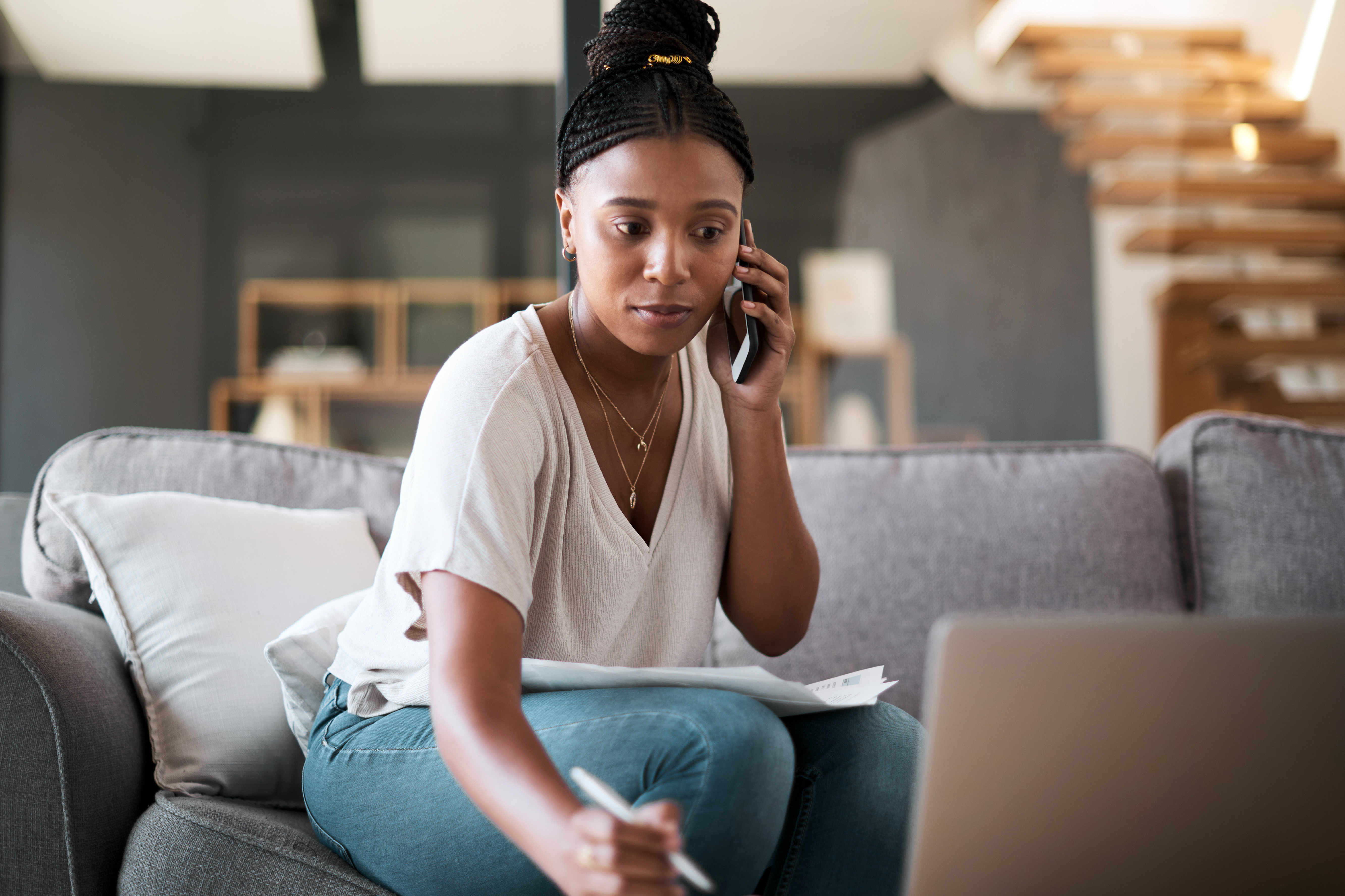A woman sitting on a couch talks on the phone while reviewing papers and taking notes on her laptop.