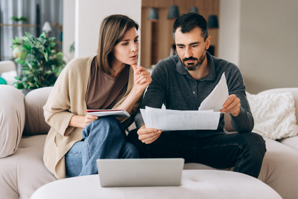 Man and woman reviewing financial documents together while sitting on a couch with a laptop nearby.