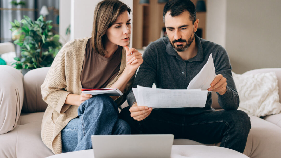 Man and woman reviewing financial documents together while sitting on a couch with a laptop nearby.