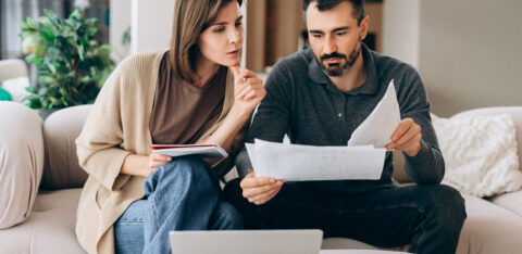Man and woman reviewing financial documents together while sitting on a couch with a laptop nearby.