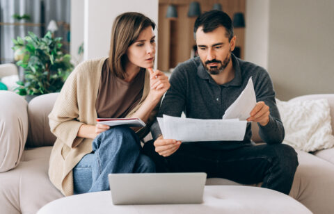 Man and woman reviewing financial documents together while sitting on a couch with a laptop nearby.