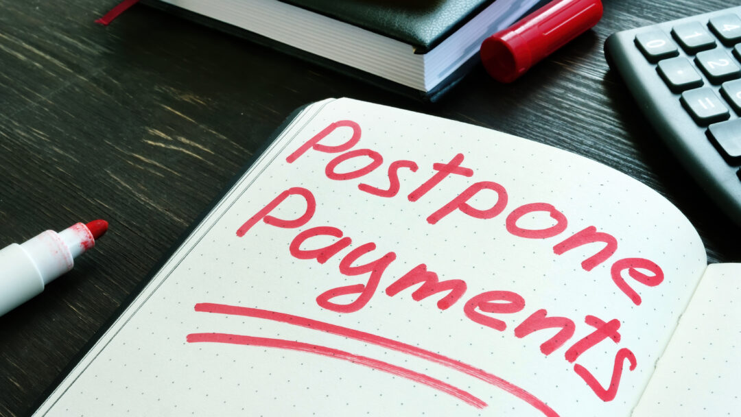 The words “postpone payments” written in red marker on an open notebook beside a calculator and books.