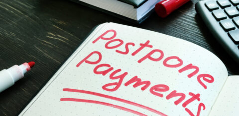 The words “postpone payments” written in red marker on an open notebook beside a calculator and books.