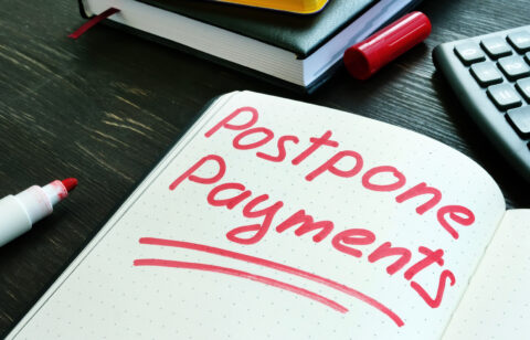 The words “postpone payments” written in red marker on an open notebook beside a calculator and books.