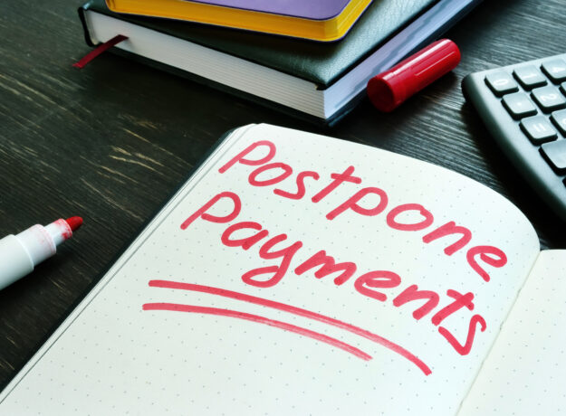 The words “postpone payments” written in red marker on an open notebook beside a calculator and books.