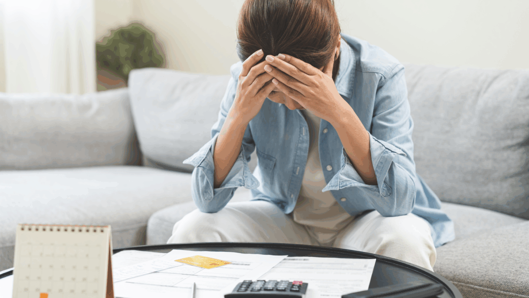 A woman sits at her coffee table with bills and a calculator, holding her head in stress while reviewing her finances.
