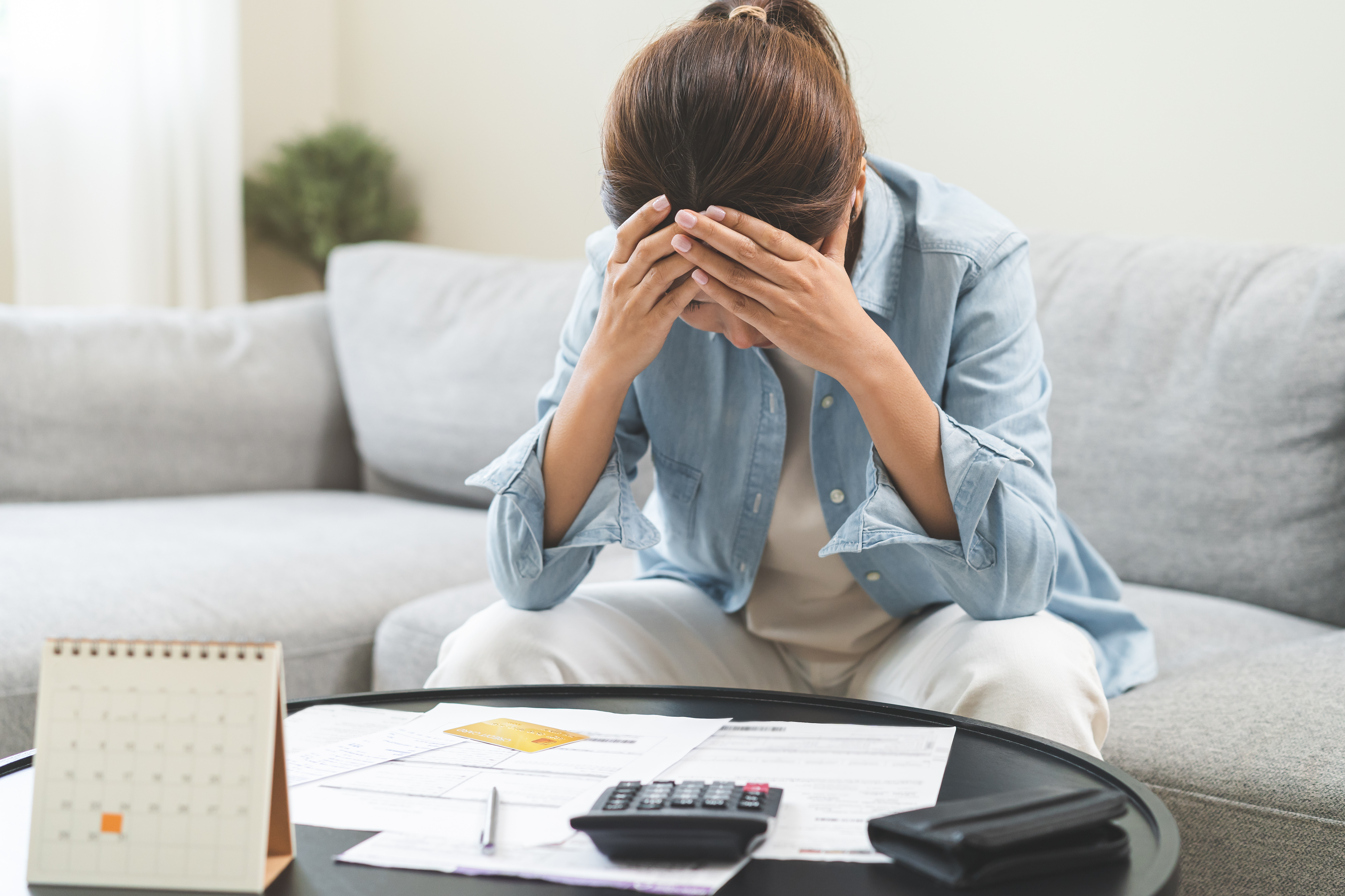 A woman sits at her coffee table with bills and a calculator, holding her head in stress while reviewing her finances.