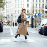 A woman wearing a long coat and backpack crosses a city street while holding a smartphone and a coffee.