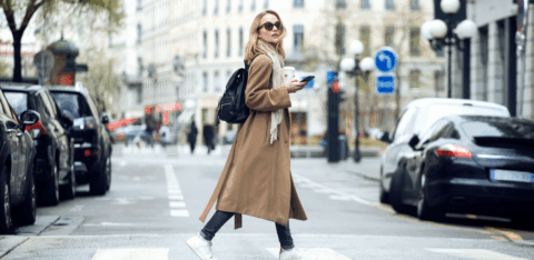 A woman wearing a long coat and backpack crosses a city street while holding a smartphone and a coffee.
